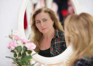 blonde vrouw kijkt in grote ronde spiegel naar zichzelf met een klein bosje roze rozen