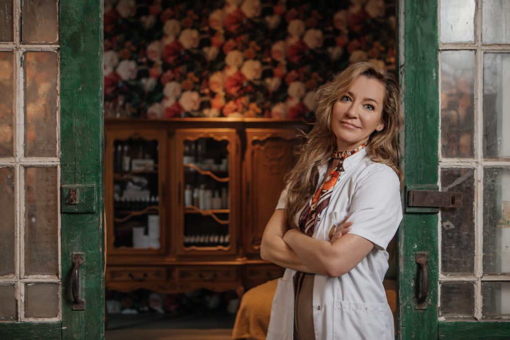 Blonde vrouw in witte blouse met das leunt tegen geopende groene deuren voor interieur studio