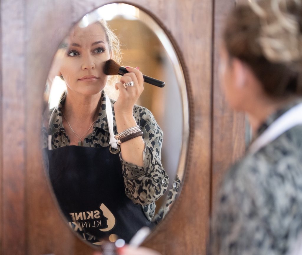 Blonde vrouw kijkt naar zichzelf in ronde spiegel in houten muur en streelt haar eigen gezicht met een make up kwast